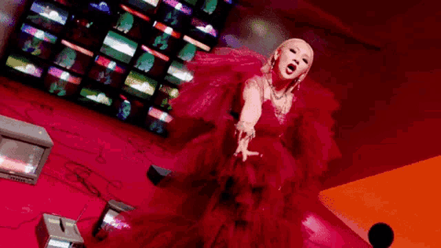 a woman in a red dress is standing in front of a wall of televisions and making a funny face .