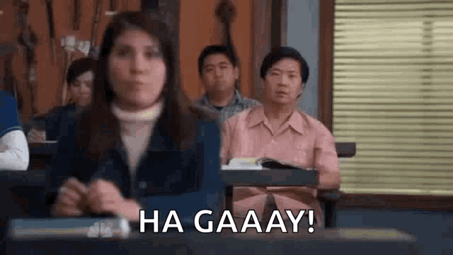 a woman is sitting at a desk in a classroom with other people and says `` ha gaaay ! ''