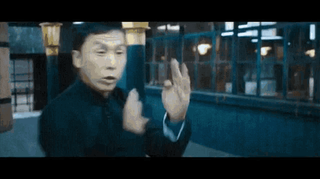 a man in a black suit is standing in front of a building with his hands up in the air .