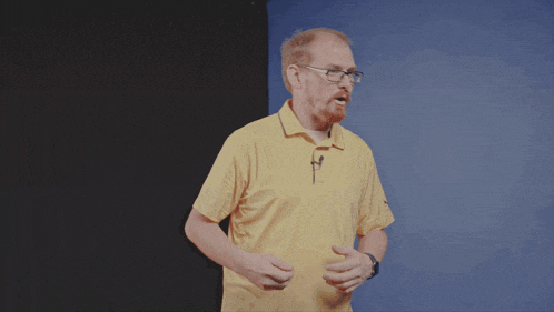 a man wearing glasses and a yellow shirt stands in front of a blue backdrop