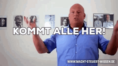 a bald man in a blue shirt is standing in front of a wall with pictures and says " kommt alle her "