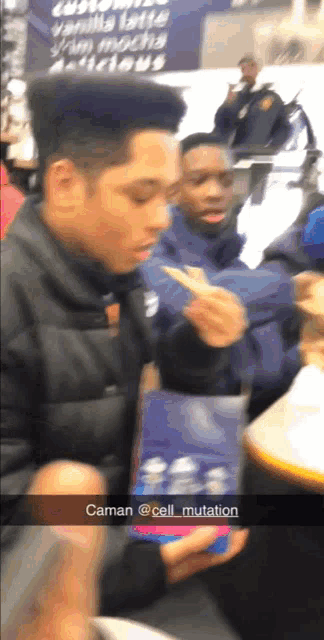 a man in a black jacket is holding a cell phone in front of a sign that says " vanilla latte "