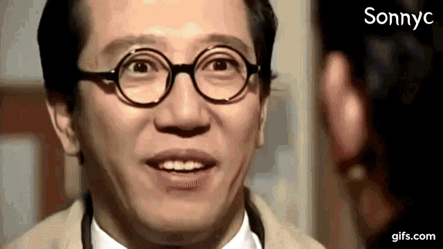 a man wearing glasses and a tan jacket is smiling and looking at another man .