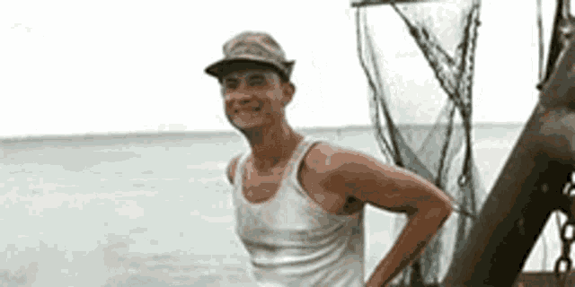 a man wearing a hat and a tank top is standing on a boat near the ocean .