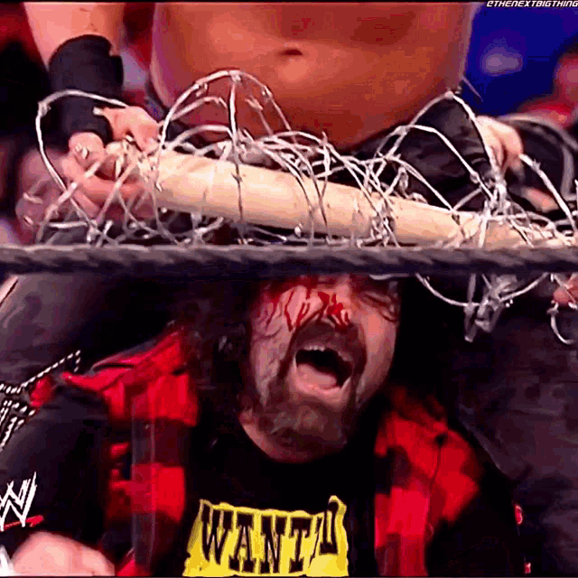 a wrestler wearing a wanted shirt holds a barbed wire fence over his head
