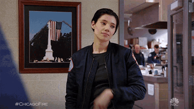 a woman in a bomber jacket is standing in front of a framed picture of an american flag on the wall ..