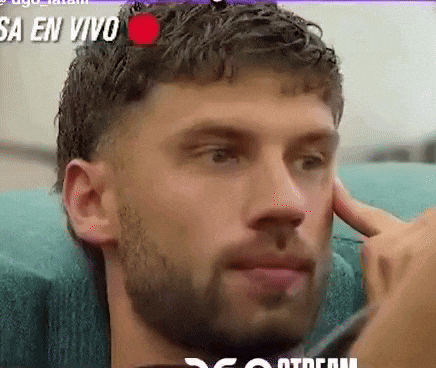 a man with a beard is sitting on a blue couch and touching his ear .