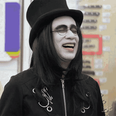 a man with long black hair and glasses is wearing a top hat and laughing