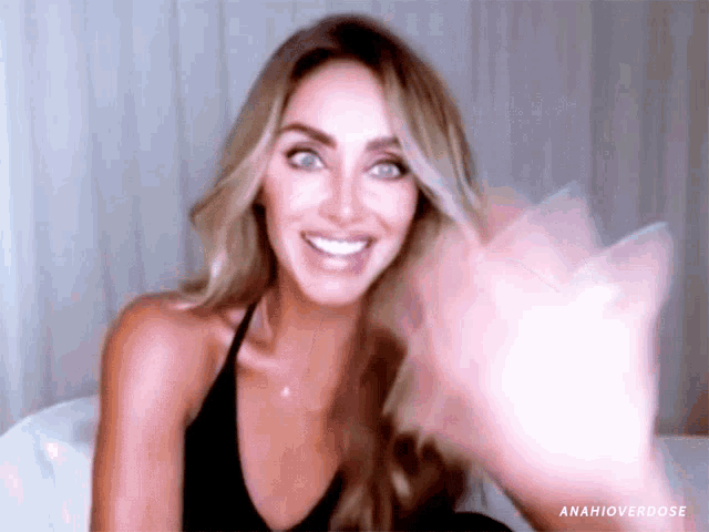 a woman in a black tank top is smiling and waving her hand