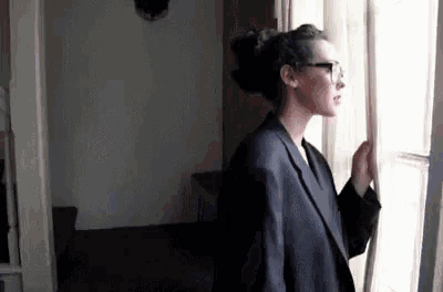 a woman in a suit and glasses is standing in front of a window looking out .