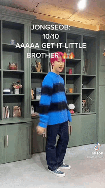 a young man in a blue and black striped sweater is standing in front of a bookshelf and says jongseob