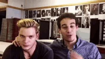 two young men are sitting next to each other in a room with pictures on the wall behind them .