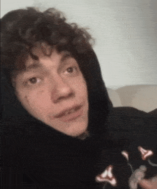 a young man with curly hair is wearing a black hoodie and making a face .