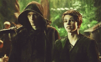 two men are standing next to each other in a forest . one of the men is wearing a hooded cape .