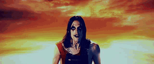 a man with long hair and black makeup stands in front of a sunset