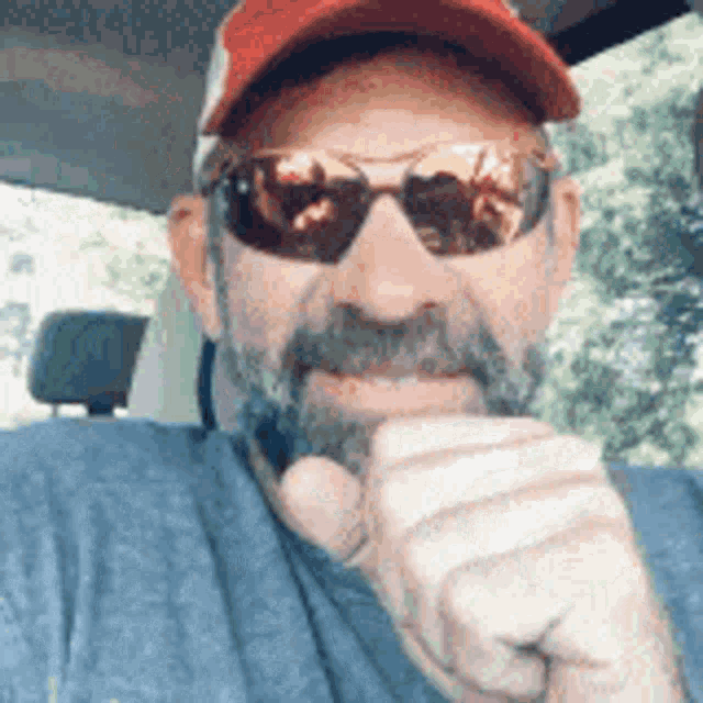 a man with a beard wearing sunglasses and a baseball cap is sitting in a car .