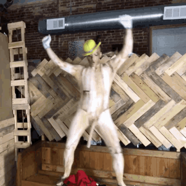 a naked man wearing a yellow hat is dancing in front of a wooden wall