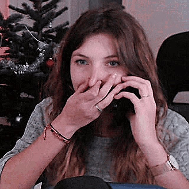 a woman covering her mouth with her hands while talking on a cell phone in front of a christmas tree