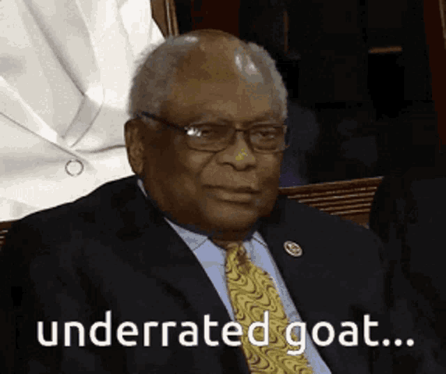 a man in a suit and tie is sitting in front of a sign that says underrated goat