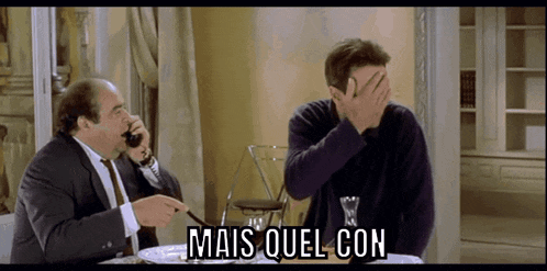 two men are sitting at a table and one of them is covering his face with his hand and the words mais quel con are written below him