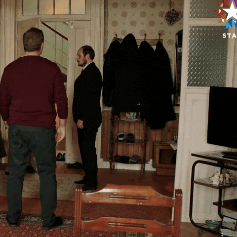a man in a suit stands in a living room next to a man in a red sweater