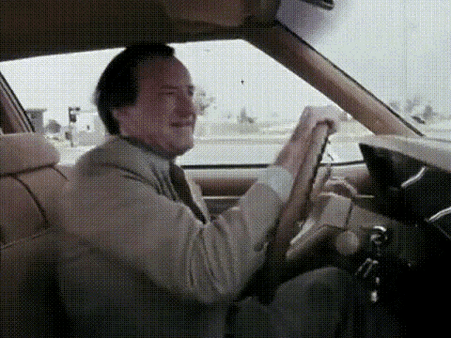 a man in a suit and tie is driving a car while holding the steering wheel .