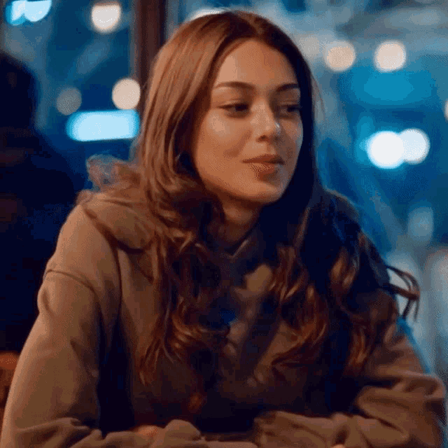 a woman in a hoodie is sitting at a table with her hands folded and looking at the camera .