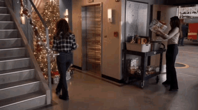 two women are standing in front of a staircase with a christmas tree and an elevator in the background