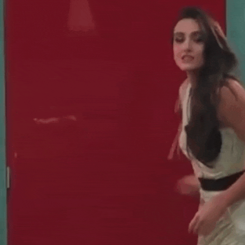 a woman in a white dress is standing in front of a red door and waving her hand .