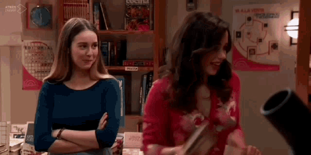 two women are standing next to each other in a room with a bookshelf .