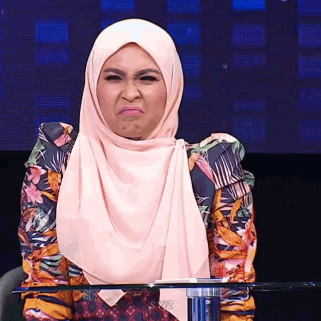 a woman wearing a hijab makes a funny face while sitting at a table