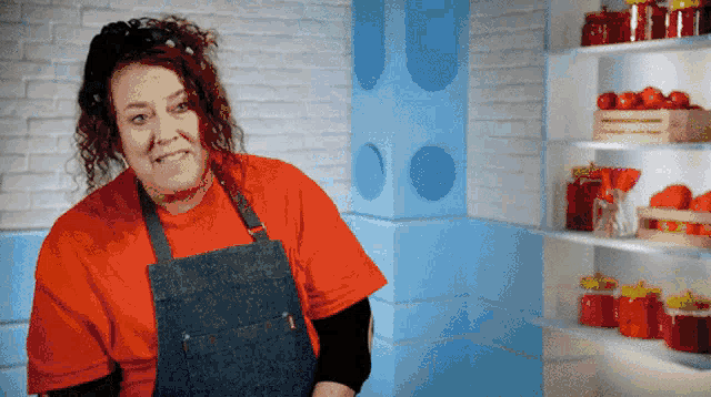 a woman wearing an apron and a red shirt stands in front of a shelf full of jars
