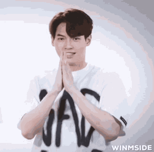 a young man in a white t-shirt is making a funny face with his hands together .