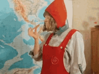 a person wearing red overalls and a red hat eating a piece of pizza in front of a map