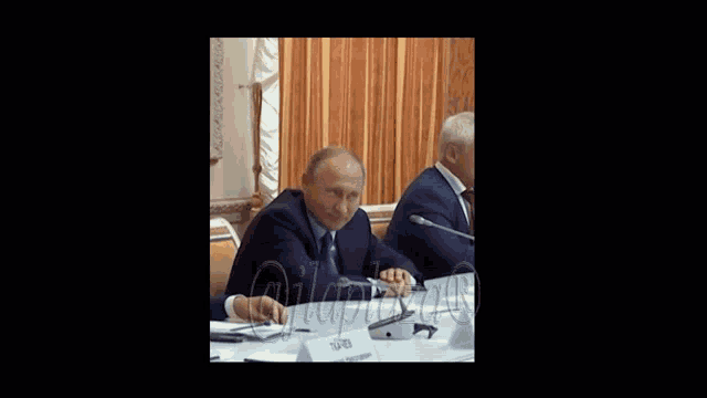 a man in a suit sits at a table with a name tag that says " putin "