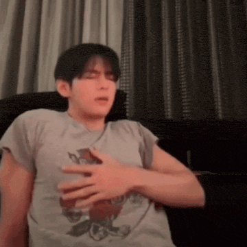 a man in a grey shirt is sitting on a couch with his hand on his chest .