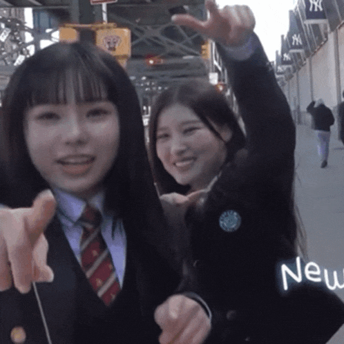 two girls are standing on a sidewalk and one of them is pointing at the camera