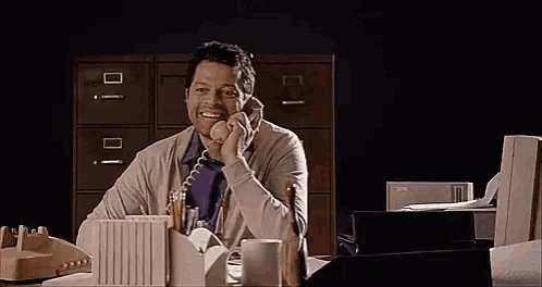 a man is sitting at a desk in an office talking on a telephone .