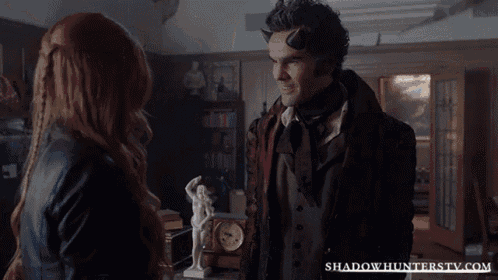 a man and a woman are standing in a living room with shadowhunterstv.com written on the bottom of the screen