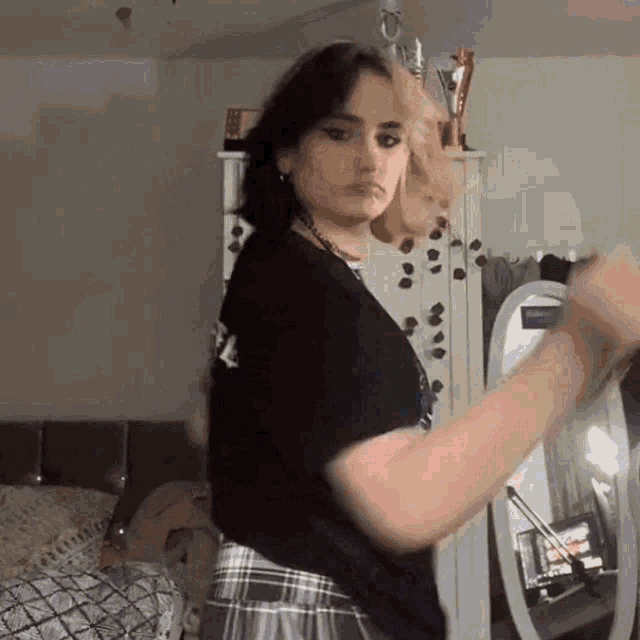 a woman in a black shirt and plaid skirt is standing in front of a mirror in a bedroom .