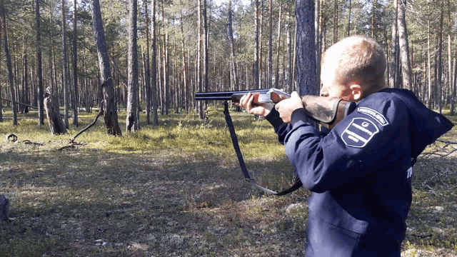 a boy is holding a shotgun in the woods and has a patch on his jacket that says ' army ' on it