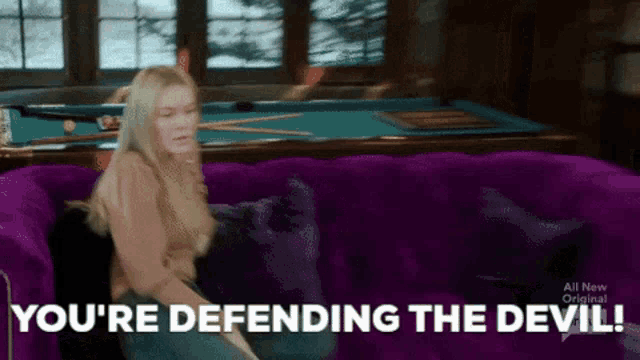 a woman is sitting on a purple couch with the words you 're defending the devil behind her