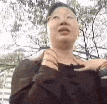 a woman wearing glasses and a black sweater is standing in front of a tree .