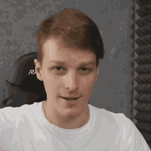 a young man in a white t-shirt is sitting in a chair and making a funny face .