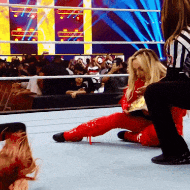 a woman in a red outfit sits on the ground in a wrestling ring while a referee looks on