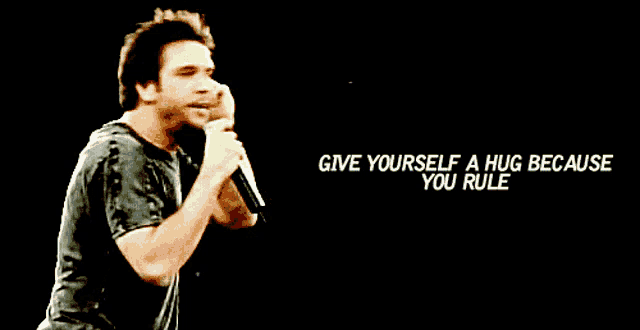 a man singing into a microphone with the words " give yourself a hug because you rule " behind him