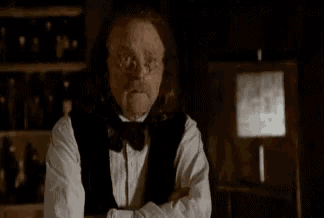 a man with long hair and glasses is standing in a dark room with his arms crossed and looking at the camera .