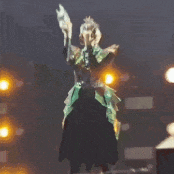 a woman in a green dress is singing into a microphone with the words okaaa written on the bottom of the image .