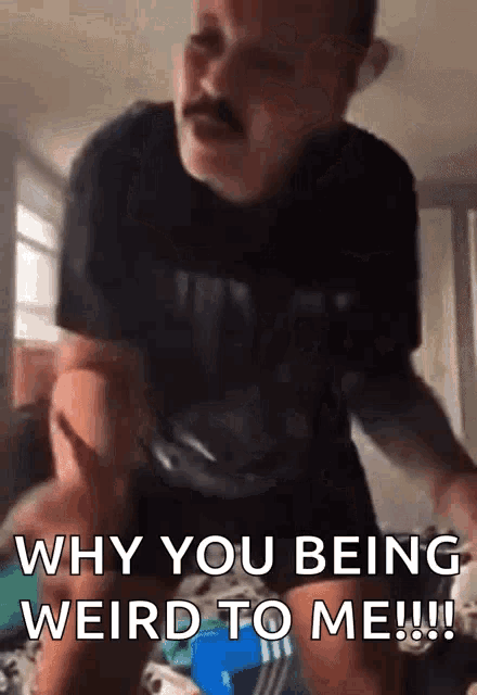 a man with a mustache is sitting on a bed and says why you being weird to me !