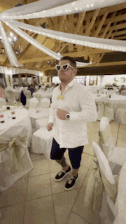 a man in a white shirt and sunglasses is dancing in a room filled with tables and chairs .